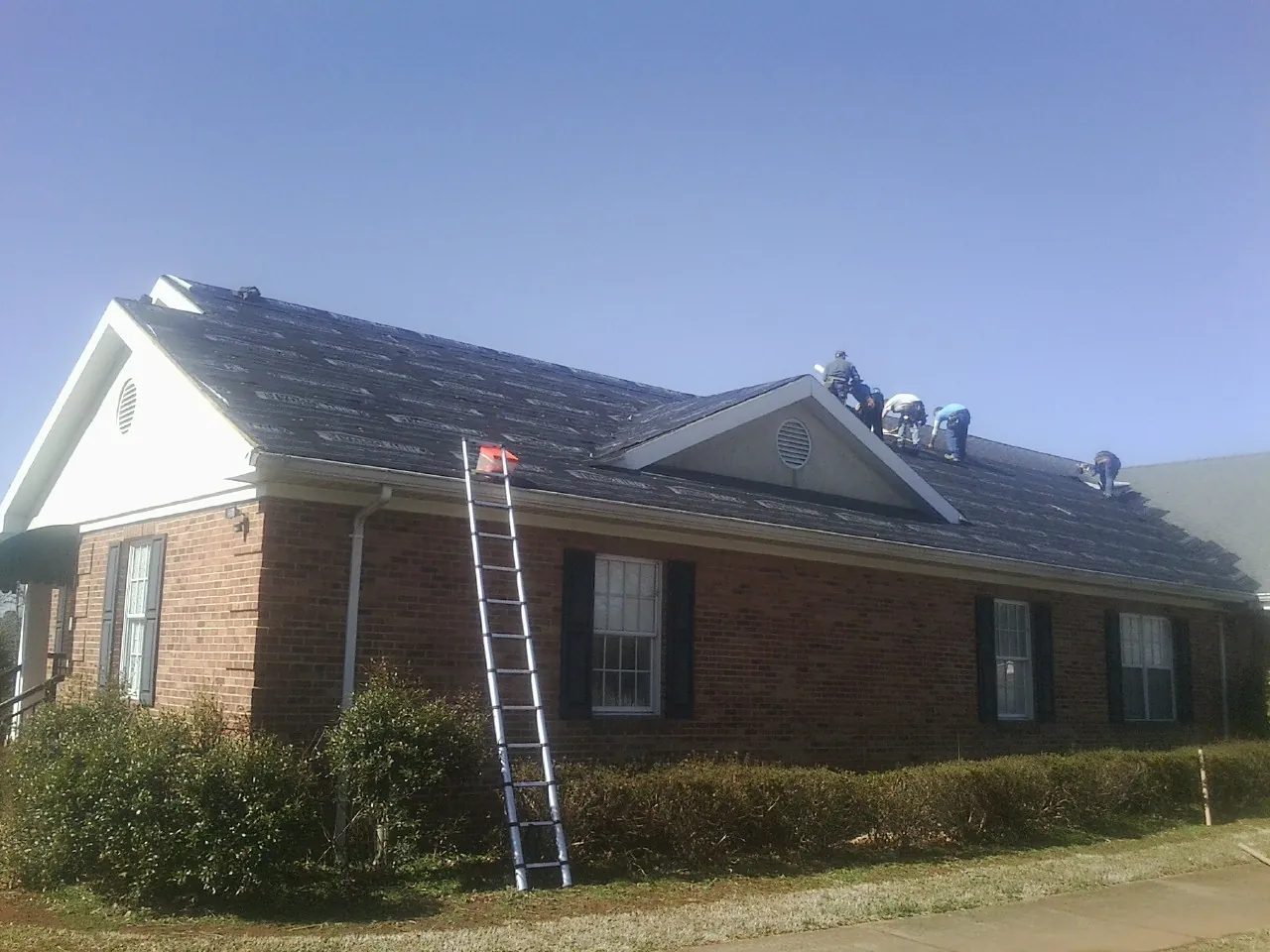 Crew tearing shingles off a brick ranch roof with a ladder set — Emergency Roof Repair work by our crew in Maxton