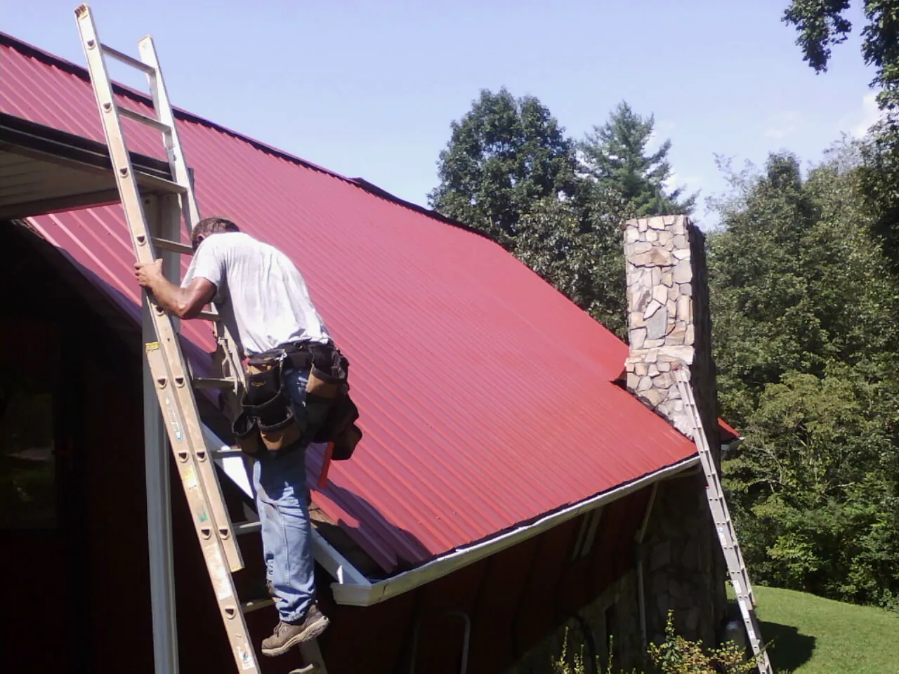 Roofer on a ladder beside a red metal roof and stone chimney — Soffit & Fascia Repair by Roofing Brothers in Maxton